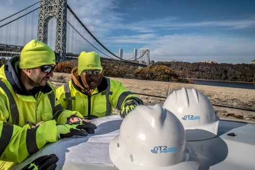 Kiwa T2UE engineers in high-visibility jackets and helmets reviewing plans near a bridge on a clear day