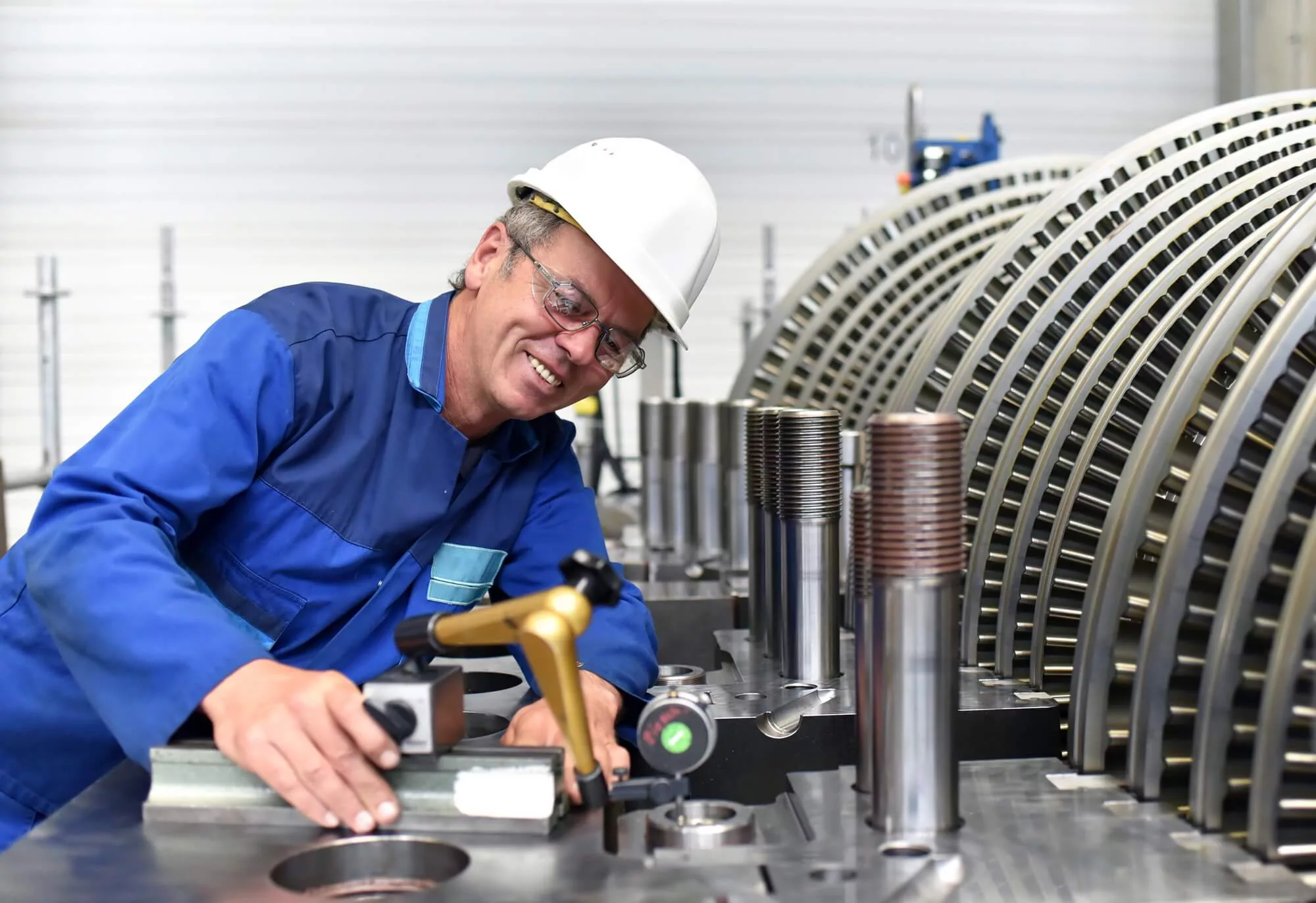A Kiwa engineer in a blue uniform and white helmet operates machinery in an industrial setting. Metal components and equipment are visible in the background.