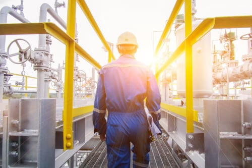Kiwa PPI engineer in blue protective gear walking through an industrial oil and gas plant platform, surrounded by pipelines and machinery, with sunlight in the background