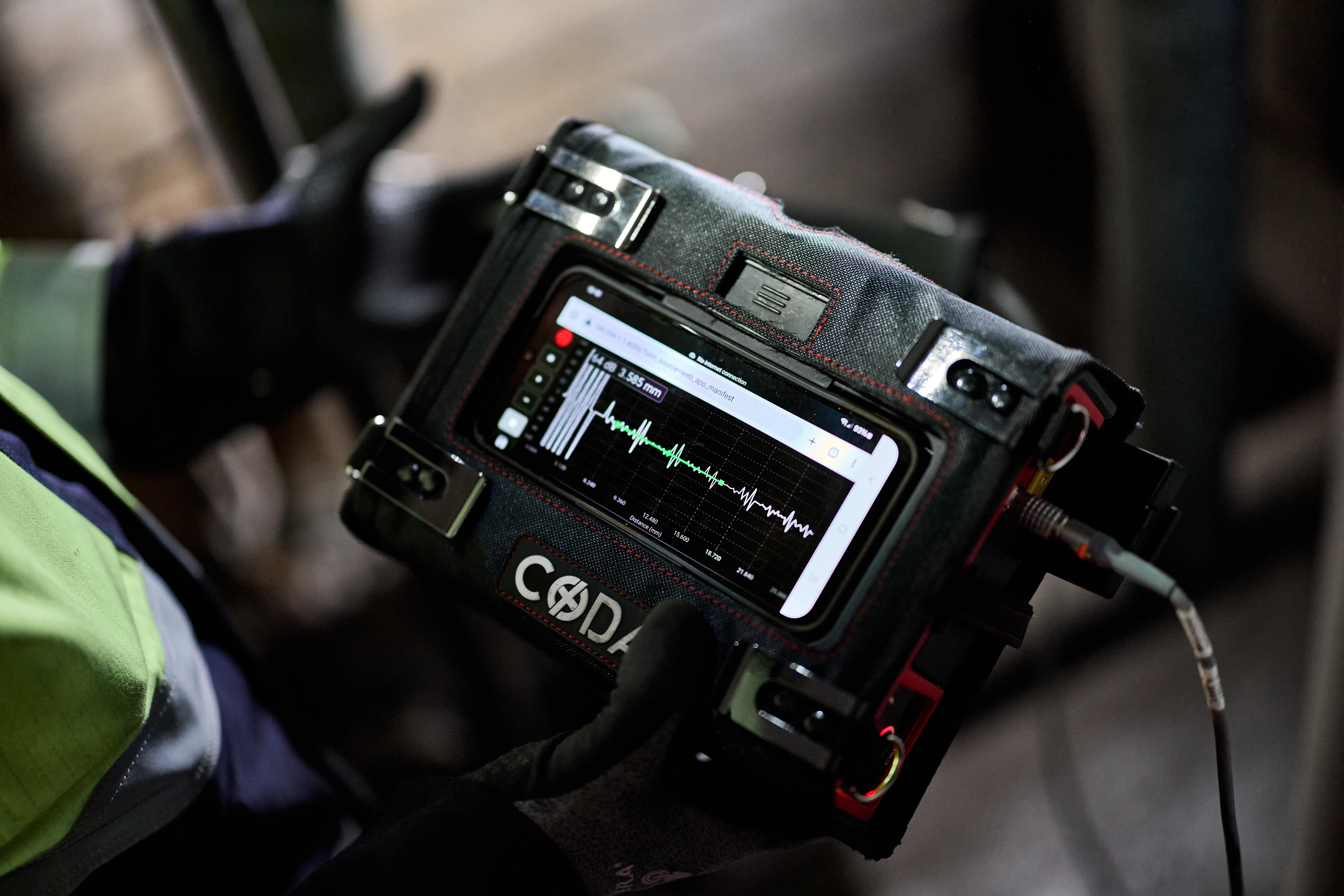 A close-up of a handheld electronic device displaying a waveform on its screen, being held by a person wearing gloves.